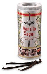 A cylindrical container of vanilla sugar stands upright, featuring a label with vanilla flowers, vanilla pods, and puddings. The text reads "Vanilla Sugar." In front of the container, three whole vanilla pods are laid out.
