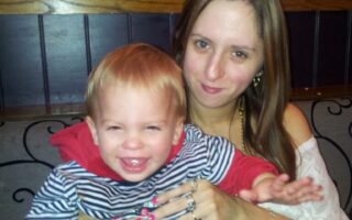 A woman with long brown hair, wearing a white top and jewellery, sits on a bench holding a smiling young child with blond hair, dressed in a red and blue striped hoodie. They appear happy and are posing closely together indoors.