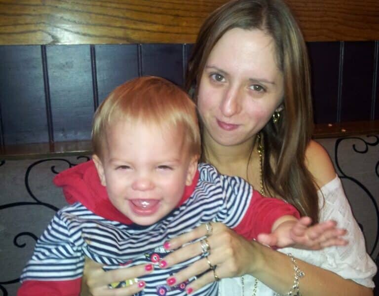 A woman with long brown hair, wearing a white top and jewellery, sits on a bench holding a smiling young child with blond hair, dressed in a red and blue striped hoodie. They appear happy and are posing closely together indoors.