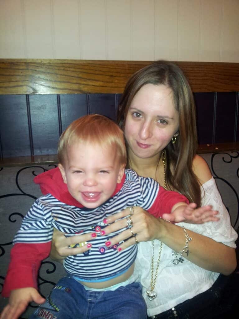 A woman with long brown hair and a white top holds a smiling toddler in her lap. The toddler has short blond hair and wears a red and blue striped hoodie. They are seated indoors against a wood-panelled and purple wall background.
