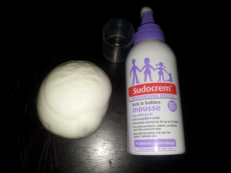 A bottle of Sudocrem kids & babies moisturising mousse is on a dark surface, with its clear cap lying nearby. Beside the bottle is a round mound of white mousse dispensed from the bottle. The bottle features purple and red labelling.