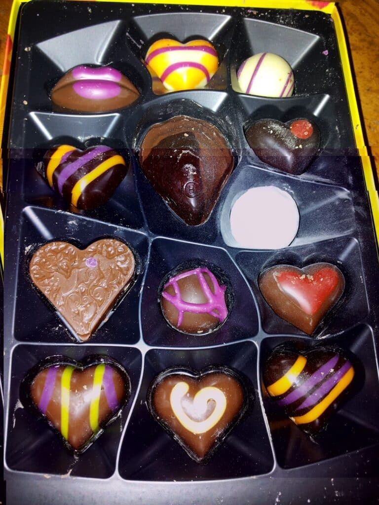 A box of assorted heart-shaped chocolates, each decorated with colourful icing in stripes, swirls, and dots of yellow, pink, orange, purple, and red. The chocolates are arranged neatly in a black plastic tray with individual compartments.