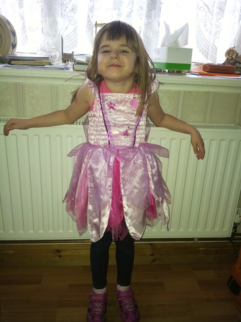 A young girl stands smiling in front of a white radiator, wearing a sparkly pink princess dress with a layered skirt. She has straight brown hair with a fringe, dark tights, and pink shoes. Behind her is a window with white curtains and various objects on the sill.