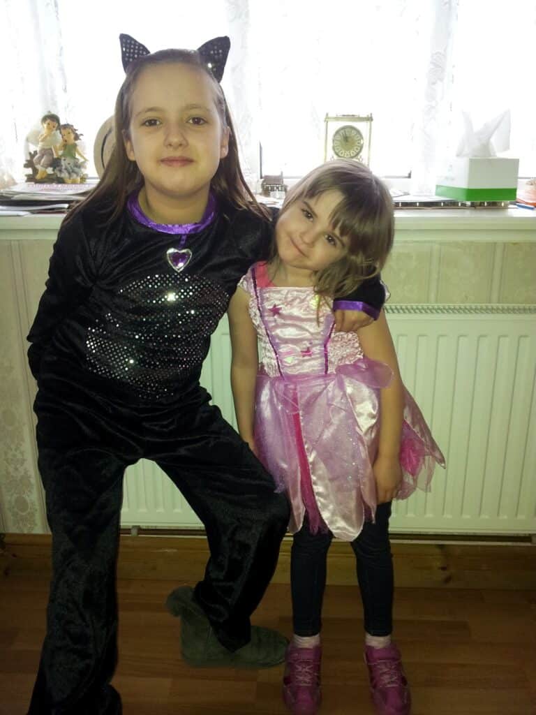 Two young girls pose indoors. The older girl wears a black cat costume with cat ears and a sparkling heart on her chest. The younger girl wears a pink fairy or princess dress with purple details and pink shoes. They stand in front of a radiator and a windowsill with ornaments.