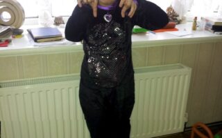 A young child stands indoors wearing a black cat costume with sequins, cat ears headband, and a purple heart necklace. The child points their fingers downward, mimicking cat paws, and smiles. Behind them is a white radiator, a windowsill with decor, and a wooden toy.