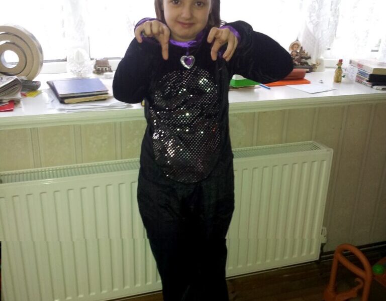A young child stands indoors wearing a black cat costume with sequins, cat ears headband, and a purple heart necklace. The child points their fingers downward, mimicking cat paws, and smiles. Behind them is a white radiator, a windowsill with decor, and a wooden toy.