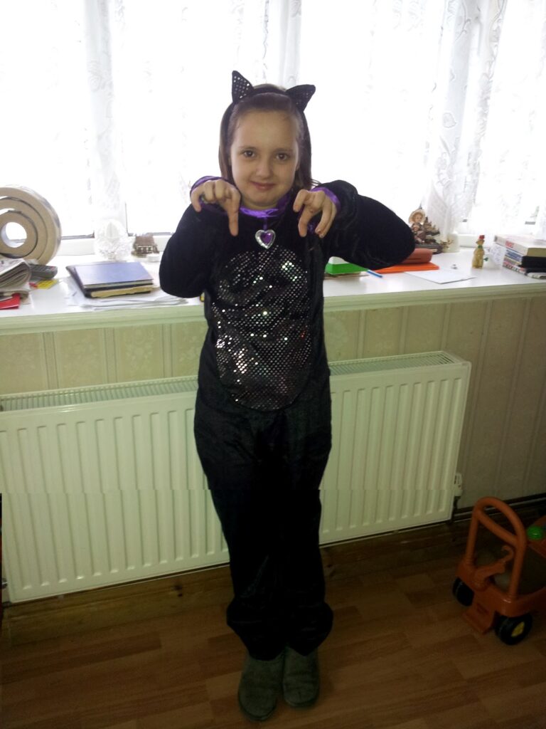 A young child stands indoors wearing a black cat costume with sparkly details, cat ears headband, and a purple heart necklace. The child is making a playful claw gesture with both hands. Sunlight shines through lace curtains behind them.