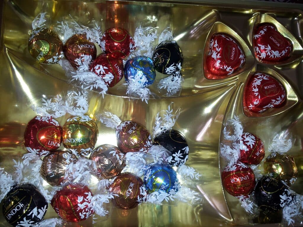 A gold tray holds assorted Lindor chocolate truffles in red, blue, black, and gold wrappers, plus two red heart-shaped Lindor chocolates in the top right section. All chocolates are wrapped in foil with white floral designs and cellophane twists.