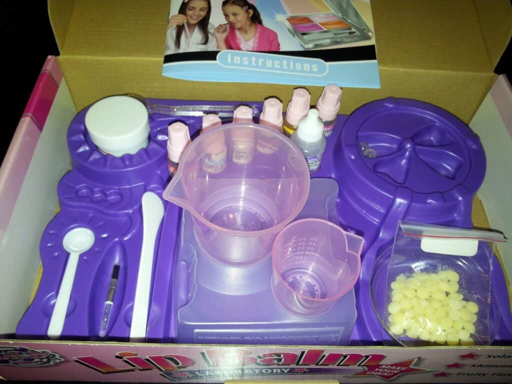 A lip balm laboratory kit in a box, featuring purple plastic moulds, measuring cups, a spoon, mixing tools, small bottles, a bag of yellow wax pellets, and an instruction booklet with images of two smiling girls on the cover, all neatly organised.