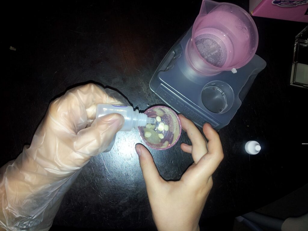 A person wearing a clear glove uses a small bottle to pour liquid into a plastic cup held by their other hand. The cup contains white pieces. A pink cup with a strainer and a grey plastic tray are also on the dark surface.
