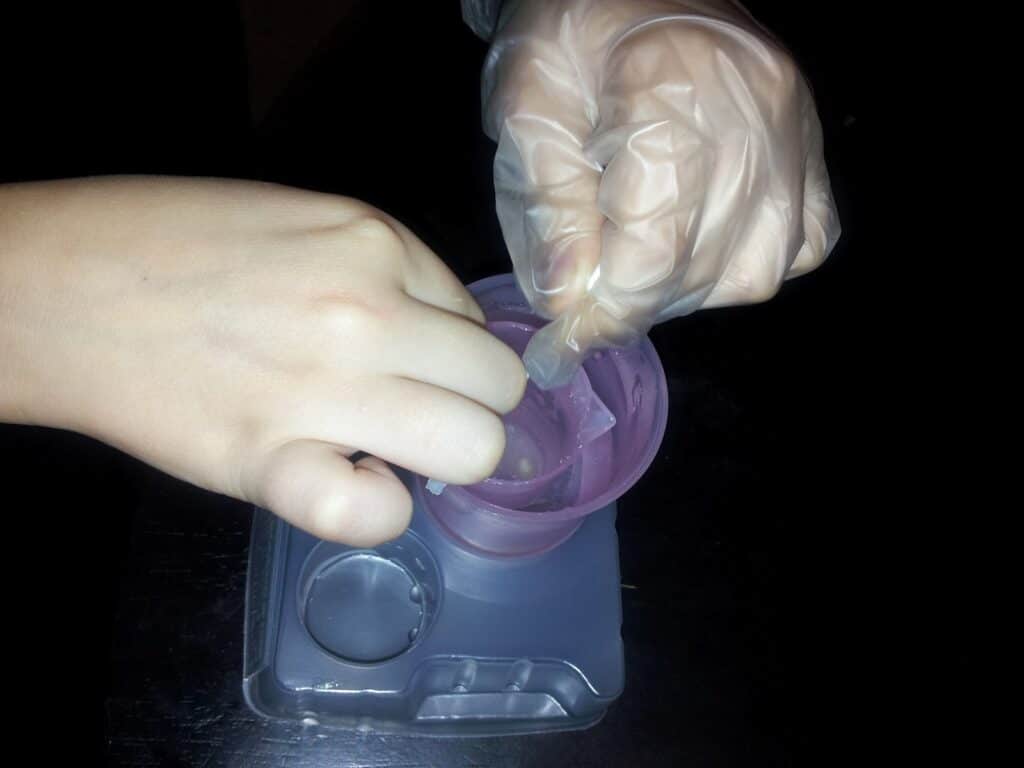A person’s hand holds a cup while another hand, wearing a disposable glove, stirs or manipulates something inside the cup. The cup sits on a small plastic tray with a circular compartment, all on a dark surface.