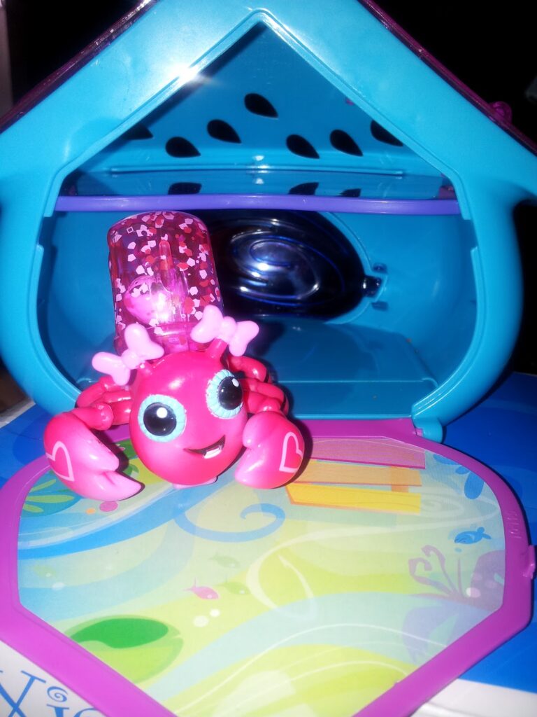 A pink toy crab with large blue eyes, heart designs, and a clear pink glittery hat sits in front of a small blue and purple plastic playhouse with an open door and a colourful patterned floor. The playhouse has heart-shaped holes on its back wall.