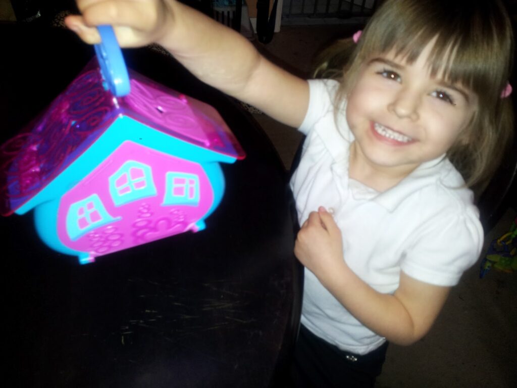 A young girl with light brown hair in plaits smiles brightly while holding a colourful toy house with a pink roof and blue base. She is wearing a white shirt and standing by a dark table. The background is softly blurred.