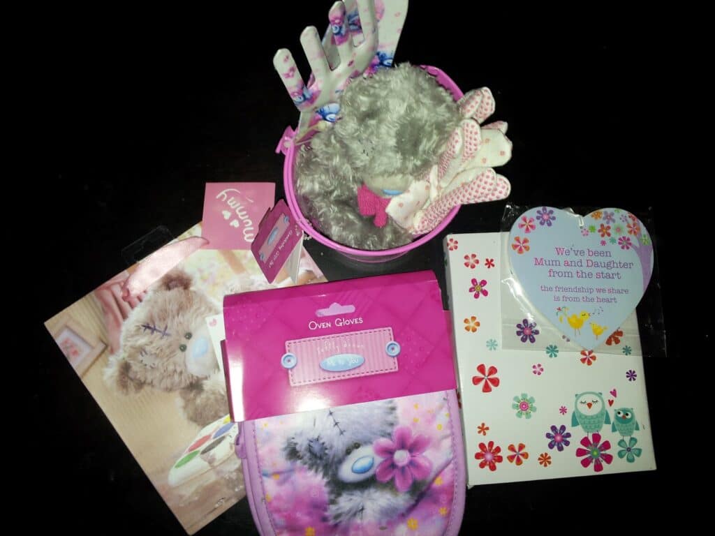A collection of mother-daughter-themed gifts on a black surface, including a plush grey teddy bear in a pink pail, floral-patterned oven gloves, a greeting card with a heartfelt message, and floral and owl-themed stickers.