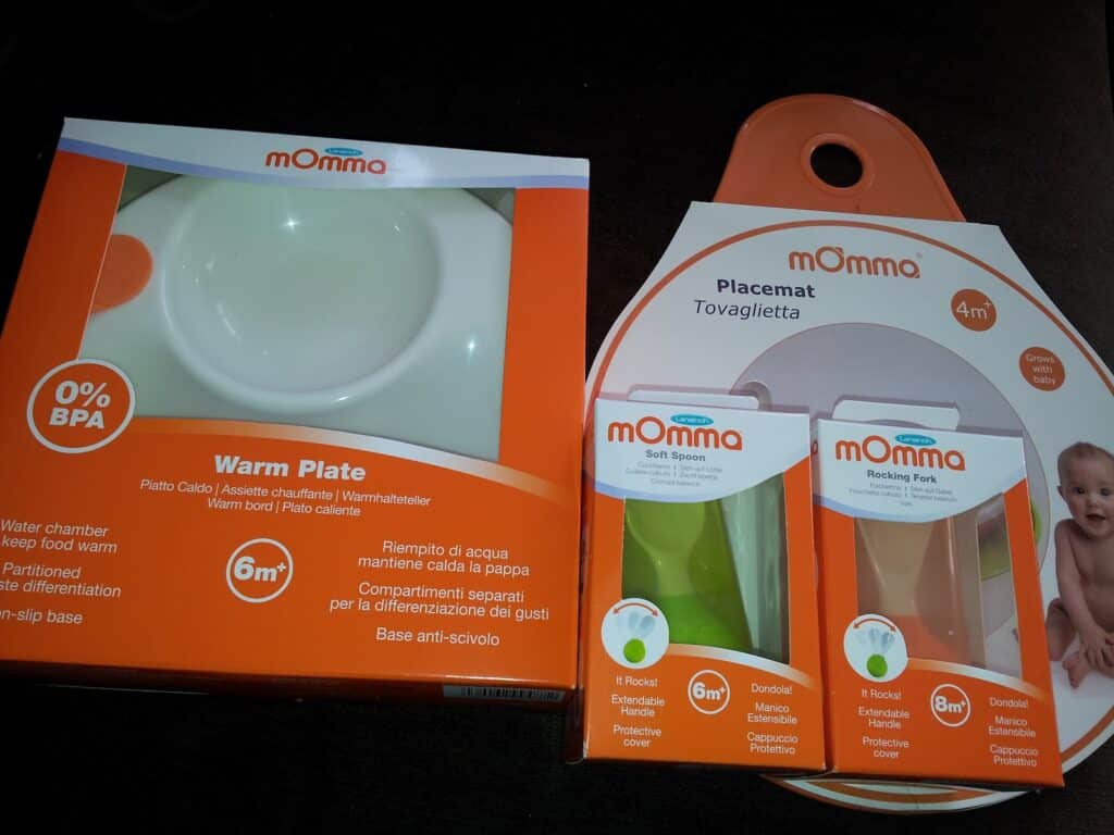 A set of baby feeding products from the brand “mOmma,” including a warm plate, a placemat, a soft spoon (6m+), and a rocking fork (12m+). The items are in orange and white packaging, with product labels and age recommendations visible.