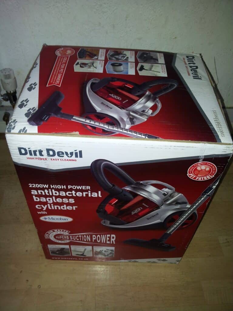 A boxed Dirt Devil vacuum cleaner is shown on a wooden floor. The box highlights features like “antibacterial bagless cylinder,” “2200W high power,” and “super suction power,” with images of the vacuum and cleaning scenes printed on the packaging.