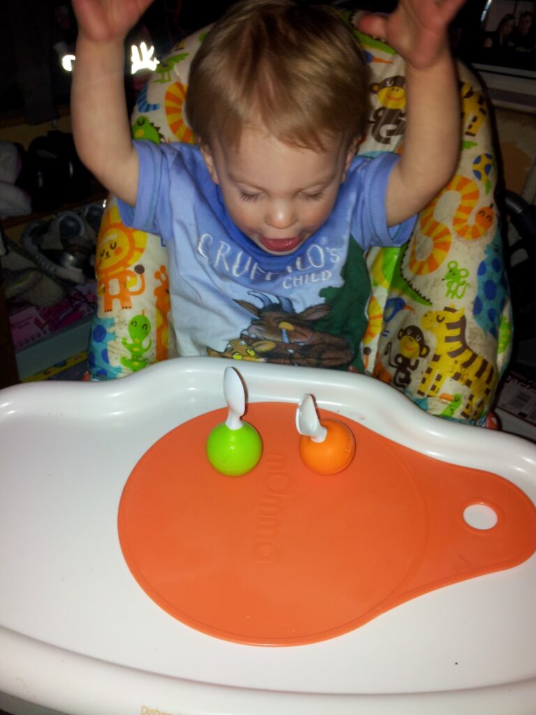 A toddler sits in a colourful highchair, raising their arms excitedly. In front of them, on an orange mat atop a white tray, are two small spoons standing upright in green and orange holders. The child is looking down at the spoons.