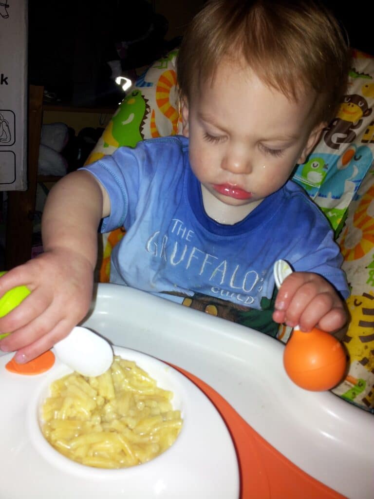 A young child with light brown hair sits in a highchair, holding a green and white spoon and an orange fork, attempting to eat macaroni cheese from a white bowl. The child wears a blue Gruffalo Club shirt and looks down at the food with concentration.