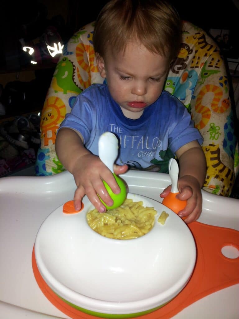 A toddler sits in a colourful highchair, focused on eating macaroni cheese from a white plate. He uses green and orange child-safe cutlery and wears a blue shirt. The background is filled with patterned fabric and baby items.