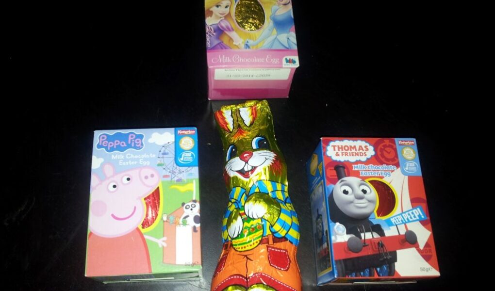 Four chocolate Easter treats on a dark surface: a pink Disney Princess egg box, a Peppa Pig egg box, a Thomas & Friends egg box, and a centre foil-wrapped chocolate bunny decorated with colourful clothes, holding a painted egg.