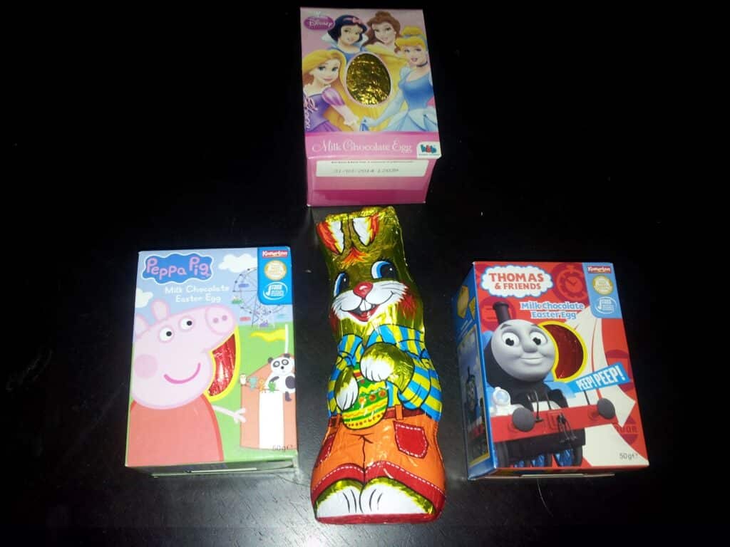 Four chocolate Easter treats on a dark surface: a Disney Princess egg at the top, a Peppa Pig egg on the left, a Thomas & Friends egg on the right, and a colourful foil-wrapped chocolate bunny standing upright in the centre.