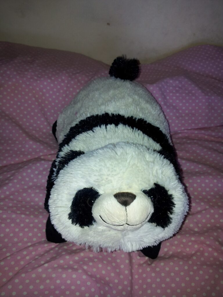 A plush panda toy with black ears, eye patches, and limbs is lying on a pink and white spotty blanket. The panda has a round face with a small brown nose and a gentle smile. The background is a light-coloured wall.