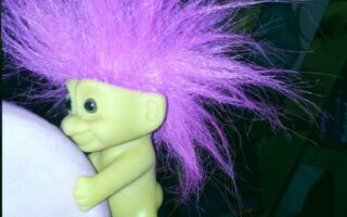 A naked troll doll with bright purple, spiky hair stands gripping the edge of a pink chair. The doll has large eyes, a wide smile, and brown-tan skin. The background is dimly lit with some indistinct furniture and dark shadows.