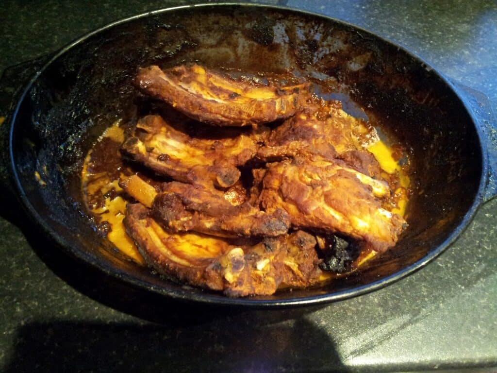 A black dish holds several cooked pork ribs coated in a dark, glossy sauce. The ribs are browned and slightly charred, with visible bone and some melted fat. The background is a dark worktop, and the dish has a rustic, homemade appearance.