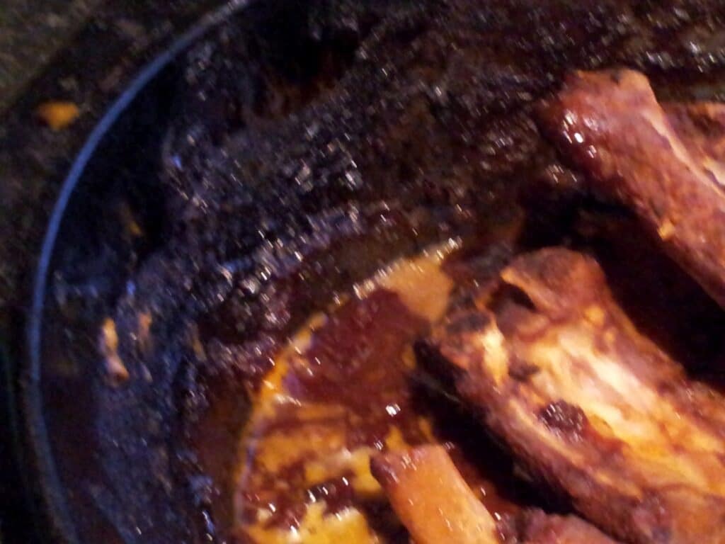 A close-up of a blackened, overcooked barbeque dish in a dark pan. Burnt sauce crusts line the pan’s sides, and partially visible ribs with charred edges sit in the remaining thick, reddish-brown sauce.