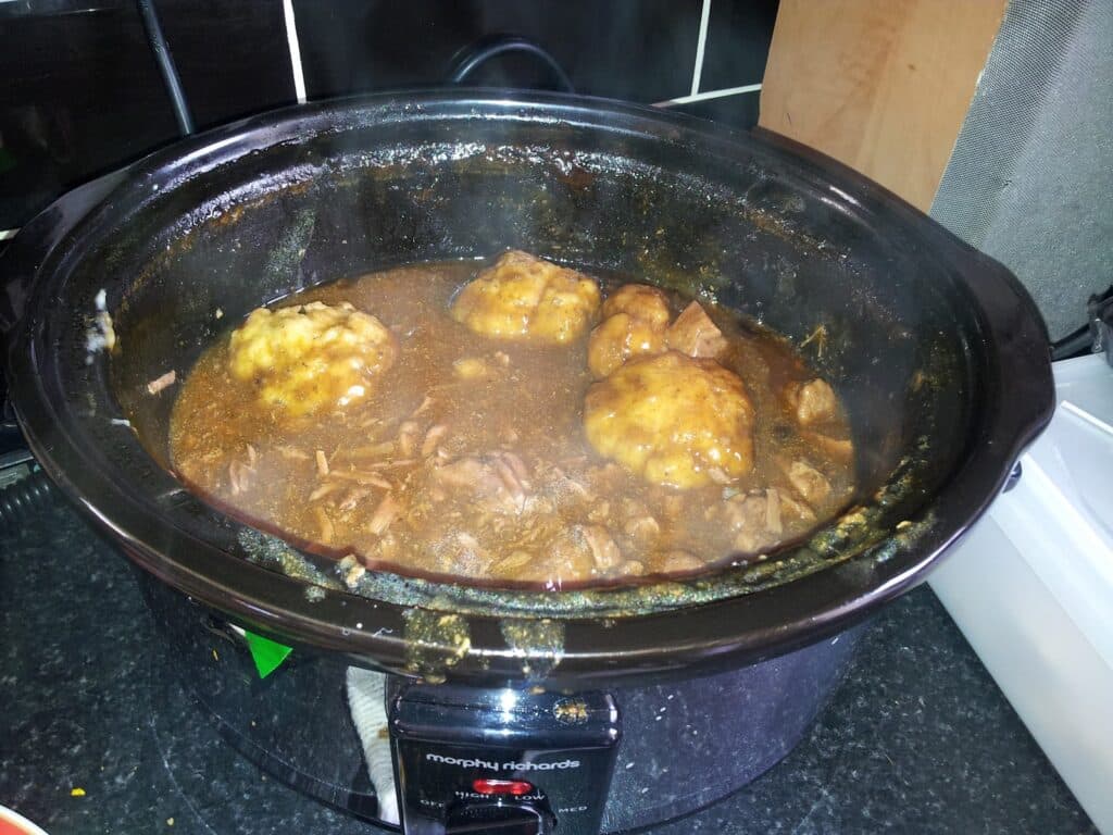 A slow cooker filled with beef stew and three dumplings in a rich brown gravy sits on a black worktop. Steam rises from the food, and chunks of meat are visible beneath the dumplings. The cooker is plugged in and switched on.