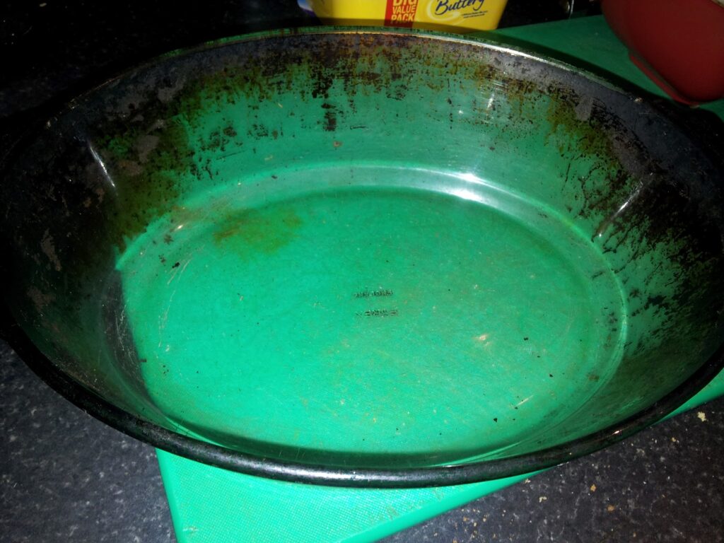 A large, round glass baking dish with dark, burnt edges and residue sits on a green chopping board atop a dark worktop. The interior of the dish is stained and shows signs of previous use. In the background, containers are partially visible.