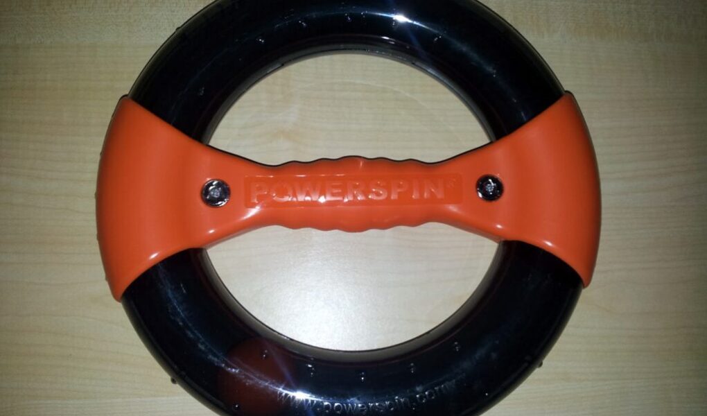 A circular exercise device with a black ring and an orange grip handle across the centre labelled "POWERSPIN," placed on a light wood surface. The handle has two visible screw heads and textured grip details.