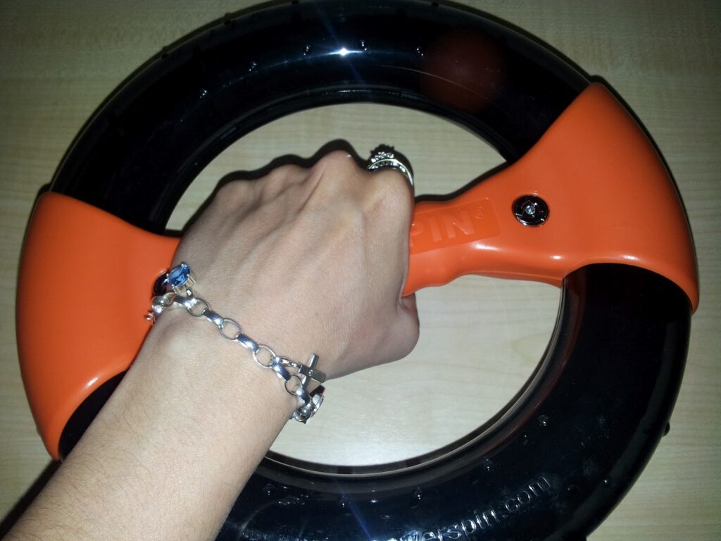 A hand wearing a silver bracelet and rings grips the orange handle of a circular black and orange handheld device, likely an exercise tool, on a light wooden surface.