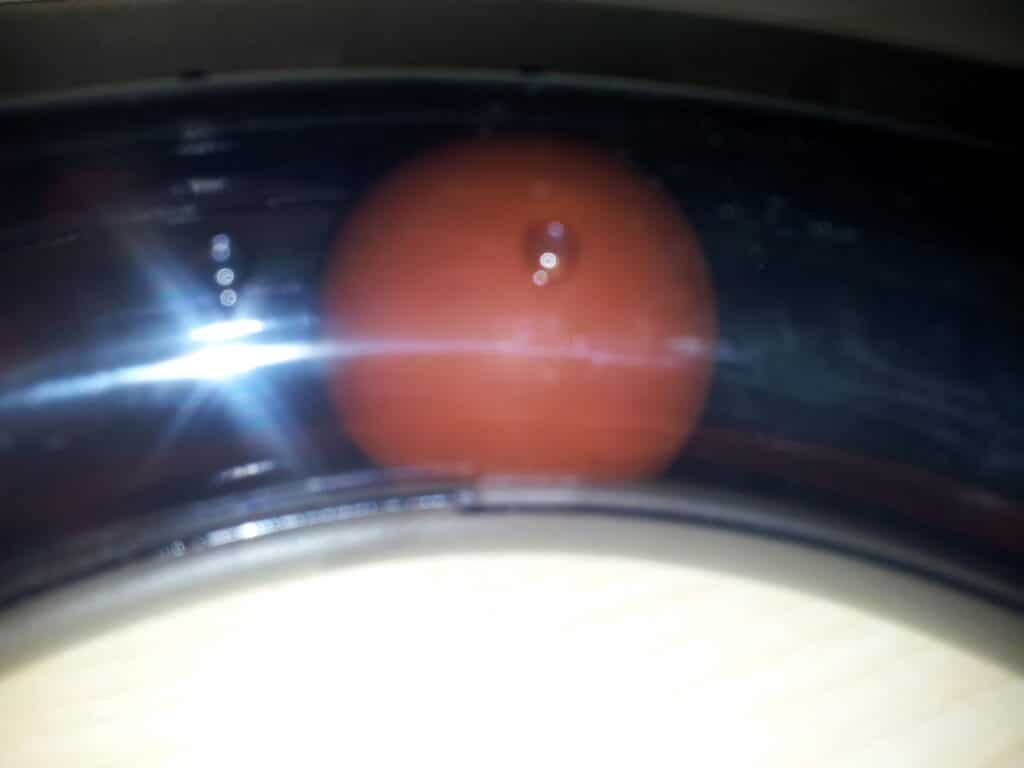 A close-up photo of a single tomato behind a transparent, curved plastic or glass surface, with a bright flash reflecting off the surface and some glare and light spots visible. The background is out of focus, creating a soft effect.
