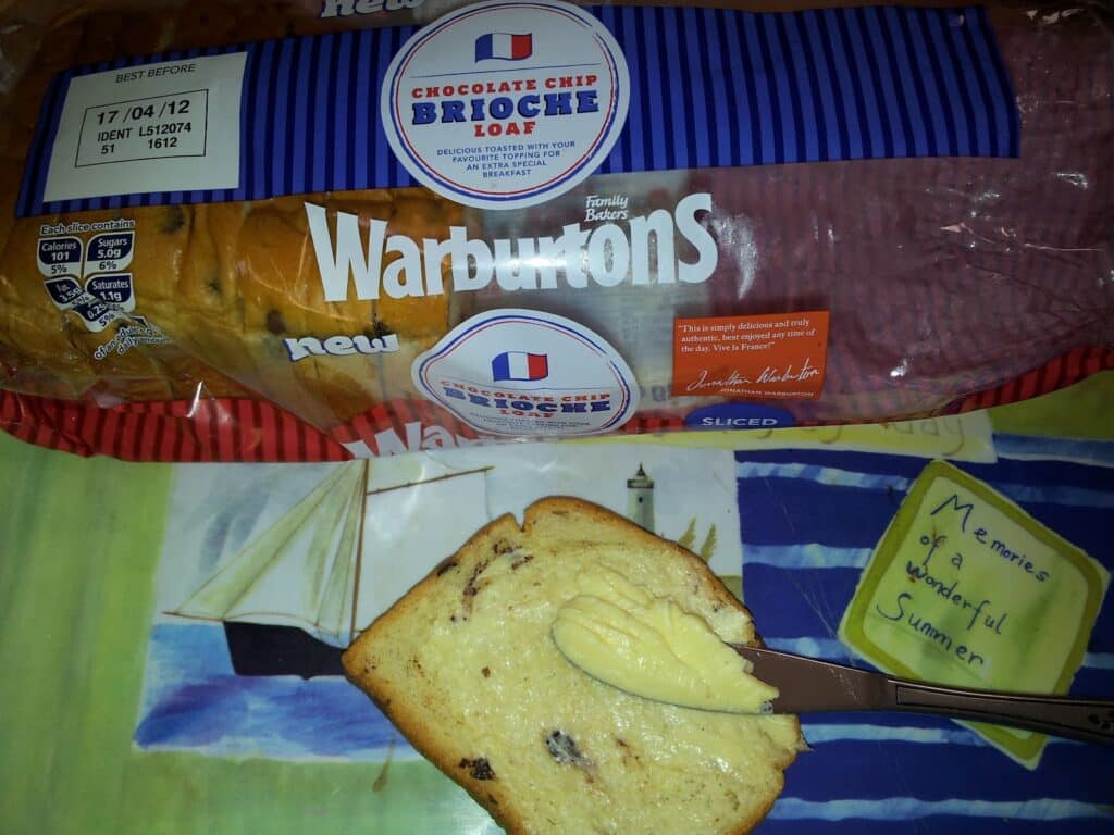 A sliced Warburtons chocolate chip brioche loaf in packaging is on a table. On a colourful plate, a slice of brioche is spread with butter by a knife. Behind the plate, there’s a yellow book titled “Memories of a Wonderful Summer.”.