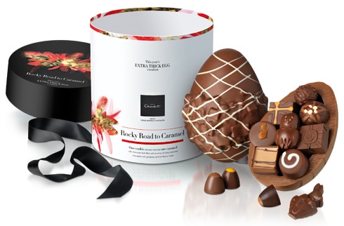 A large chocolate Easter egg, half open to reveal assorted filled chocolates inside. The egg and chocolates are displayed beside a decorative cylindrical box with floral designs, a black lid, and a black ribbon, labelled “Rocky Road to Caramel”.