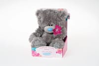 A fluffy grey teddy bear with a blue nose sits in a pink and blue floral display box. The bear has a stitched patch on its chest and holds a pink flower. The box reads, "Just for You with Love." The background is plain white.