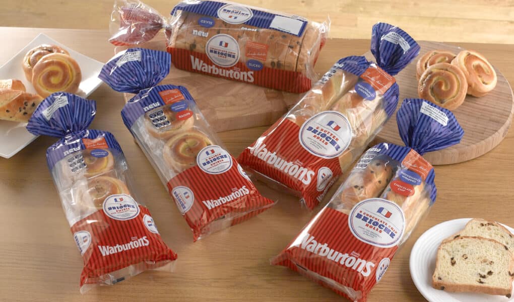 Four loaves of Warburtons bread are displayed on a wooden table, each in red, white, and blue packaging. There are also slices of bread, a white plate with a slice, and two boards with swirled pastries and sliced bread in the background.