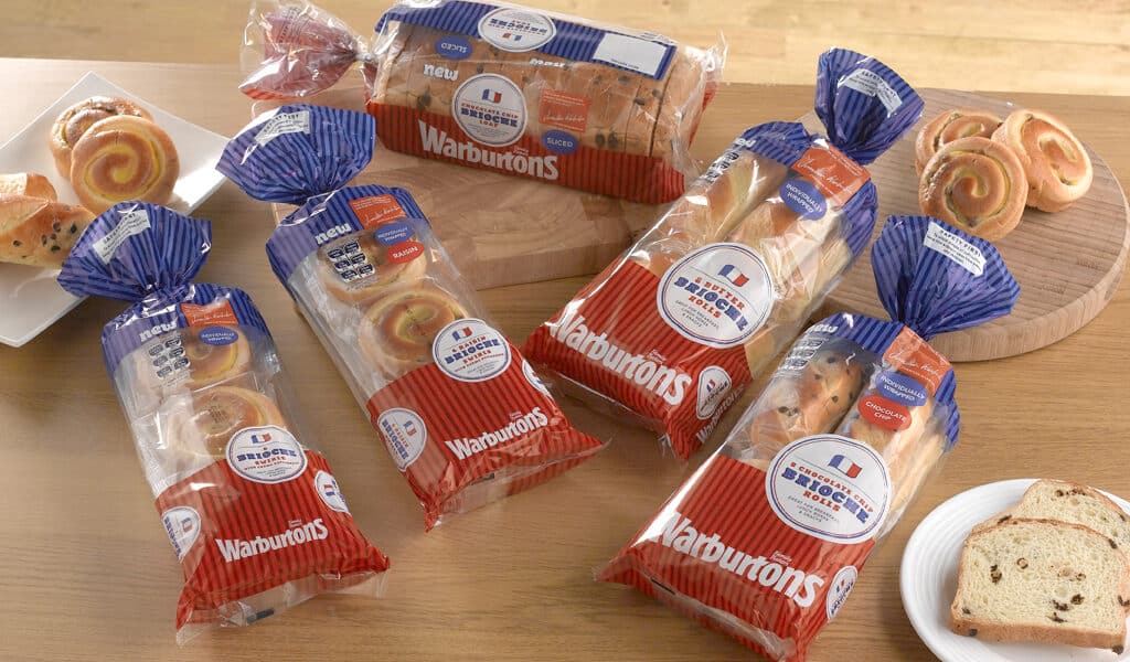 Four loaves of Warburtons bread are displayed on a wooden table, each in red, white, and blue packaging. There are also slices of bread, a white plate with a slice, and two boards with swirled pastries and sliced bread in the background.