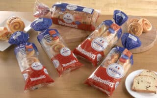 Four loaves of Warburtons bread are displayed on a wooden table, each in red, white, and blue packaging. There are also slices of bread, a white plate with a slice, and two boards with swirled pastries and sliced bread in the background.