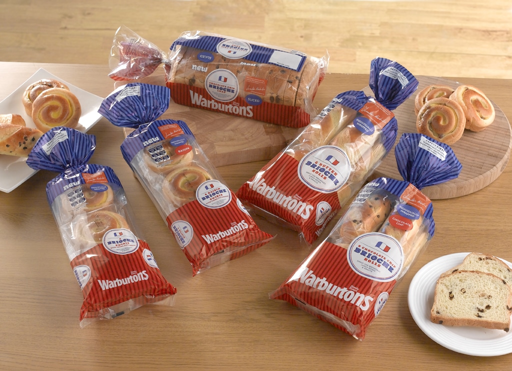 Four loaves of Warburtons bread are displayed on a wooden table, each in red, white, and blue packaging. There are also slices of bread, a white plate with a slice, and two boards with swirled pastries and sliced bread in the background.