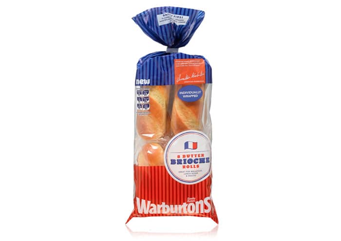 A clear plastic bag of Warburtons 6 Butter Brioche Rolls is shown. The packaging is blue, red, and white, featuring an image of brioche rolls inside and a French flag icon. The top is tied with a blue twist, and the label reads “Individually Wrapped”.