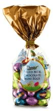A clear plastic bag filled with colourful, foil-wrapped chocolate eggs is tied at the top with shiny gold foil. The label on the front reads "Solid Milk Chocolate Mini Eggs." The assorted eggs are pink, green, blue, and gold.