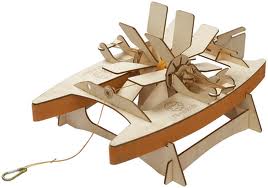 A small, wooden, toy paddle boat features two flat pontoons, a central rotating paddle wheel with blades, and a thin pull cord attached at the front. The boat has interlocking parts and a natural wood finish.