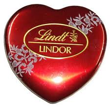 A red, heart-shaped Lindt Lindor chocolate tin with white floral designs. The tin has a shiny finish and a gold oval label in the centre with "Lindt" and "LINDOR" written in elegant script.