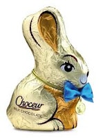 A gold-foil-wrapped milk chocolate bunny sits upright, decorated with a blue bow around its neck and a small gold medallion. The wrapper has drawn-on features: eyes, nose, mouth, and ear outlines. The label reads “Choceur MILK CHOCOLATE”.