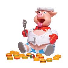 A plastic pig chef toy with a white chef's hat, red scarf, and white apron sits with a spatula in one trotter. Its mouth is open wide and it appears excited. Colourful plastic burger pieces are scattered in front of it.