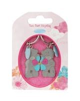 A packaged set of two grey bear keyrings is shown. Each bear holds a blue and pink flower. The background of the packaging is decorated with pastel flowers in pink, white, and blue. The label at the top reads, "Two Bear Keyrings.