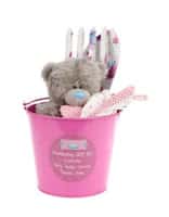 A small pink bucket holds a grey teddy bear with a blue nose, pink and white polka-dot mittens, and a pair of baby socks. The bucket has a label on the front and contains additional baby items, likely for a gift set. The background is white.