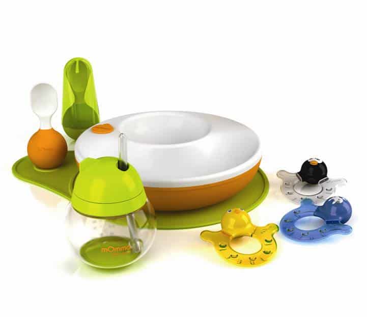 A colourful baby feeding set includes a white and orange bowl on a green mat, a green and white spoon, a clear jar with a green lid and straw, and three animal-shaped teethers in yellow, blue, and black with smiling faces and handles.